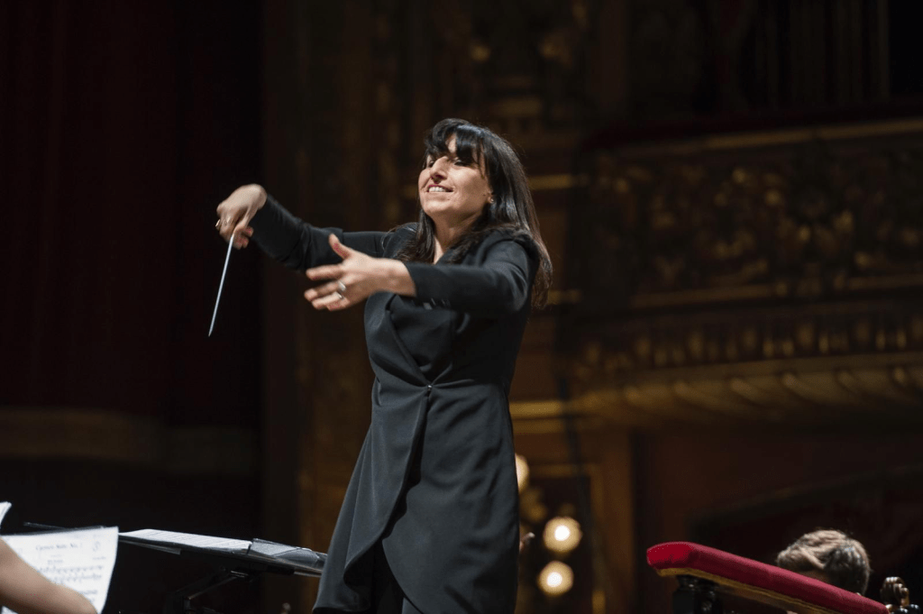 A female conductor passionately leading an orchestra on stage, with her arms raised and engaged in the music performance.